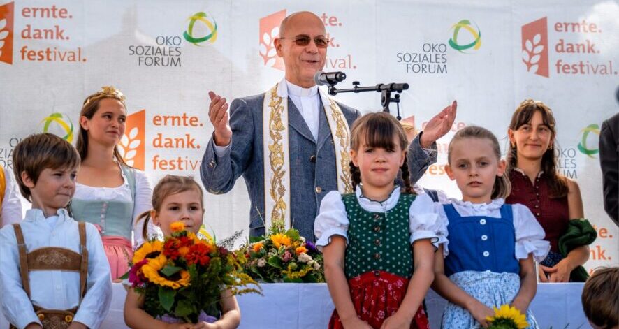 2025-09-07_EDF25_(c)ÖSF-Pöhlmann-1580-Edit Segnung beim ernte.dank.festival.: Ein Priester spricht auf der Bühne, umgeben von Kindern in Tracht mit Blumen und Erntekörben.