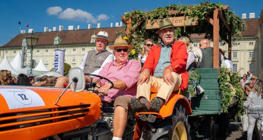 2025-09-07_EDF25_(c)ÖSF-Pöhlmann-1671 Historischer Traktor mit Anhänger beim ernte.dank.festival. am Wiener Heldenplatz, besetzt mit Männern in traditioneller Kleidung.
