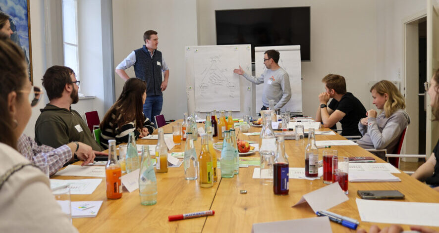 Gruppe von Teilnehmenden sitzt an einem gro&szlig;en Tisch im Seminarraum, w&auml;hrend zwei Personen vorne ein Diagramm auf einem Flipchart pr&auml;sentieren.