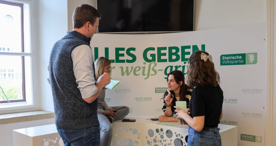 Vier Personen stehen und sitzen an einem Informationsstand in einem Innenraum und diskutieren miteinander.