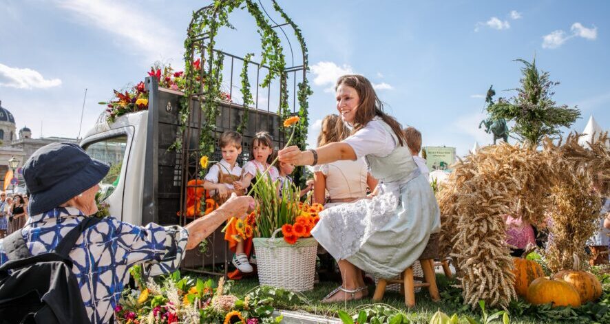 ernte.dank.festival.2025 Frau in Dirndl verteilt Blumen von einem geschmückten Erntewagen an Besucherinnen und Besucher beim Erntedankumzug am Wiener Heldenplatz