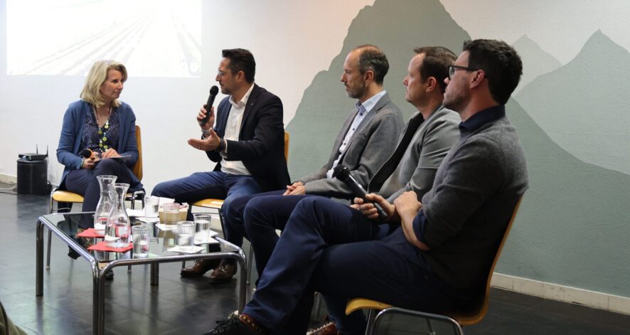 Podiumsdiskussion in einem Veranstaltungsraum: Eine Moderatorin und vier M&auml;nner sitzen nebeneinander auf St&uuml;hlen und halten Mikrofone. Ein Teilnehmer spricht gerade und gestikuliert mit der Hand, w&auml;hrend die anderen zuh&ouml;ren. Vor ihnen steht ein Tisch mit Wasserkaraffen und Gl&auml;sern, im Hintergrund ist eine projizierte Pr&auml;sentationsfolie zu sehen.