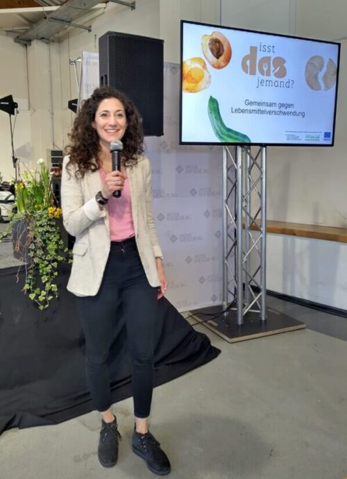 Eine Frau im Blazer steht mit einem Mikrofon vor einem Bildschirm, auf dem ein Logo mit der Aufschrift „Isst das jemand? Gemeinsam gegen Lebensmittelverschwendung!“ zu sehen ist.