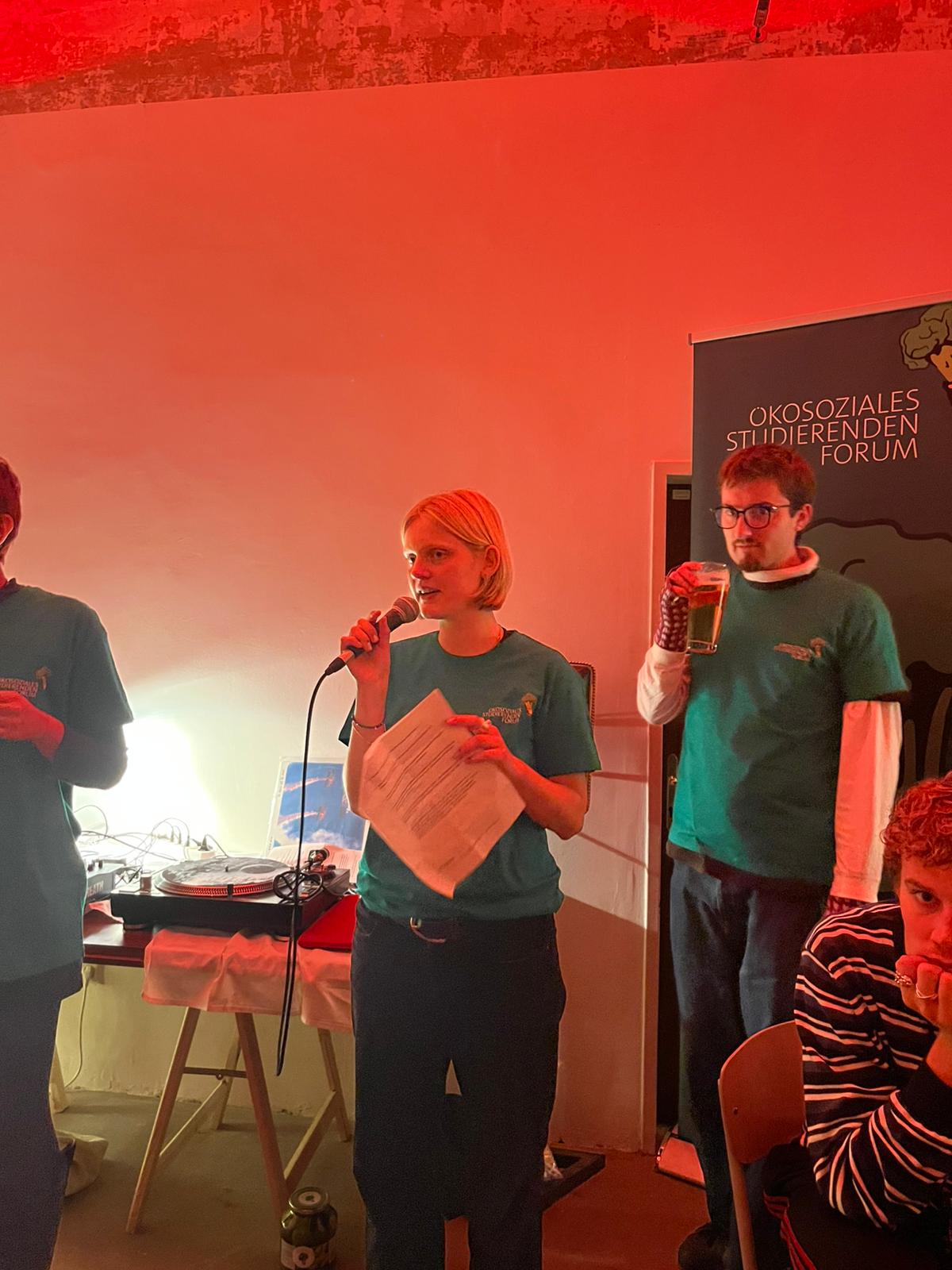 Ökosoziales Studis, 10 Pub Quiz 2026, © ÖSSFO/ Vögler Eine junge Frau moderiert eine Veranstaltung des Ökosozialen Studierendenforums und spricht mit Mikrofon vor den Teilnehmer:innen. Im Hintergrund stehen weitere Mitglieder des Organisationsteams.
