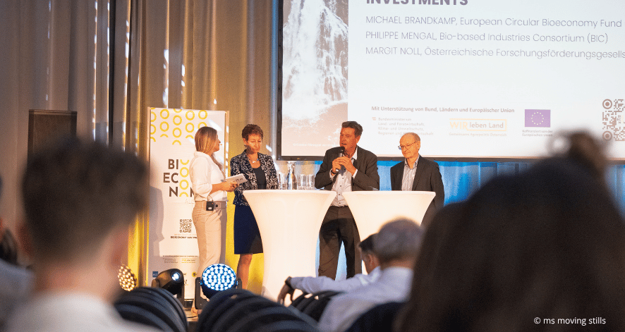 Eine Moderatorin und drei Podiumsg&auml;ste diskutieren beim Bioeconomy Summit 2026 auf einer beleuchteten B&uuml;hne. Ein Mann spricht in ein Mikrofon, w&auml;hrend die anderen zuh&ouml;ren. Im Vordergrund sind Teilnehmende aus dem Publikum zu sehen.