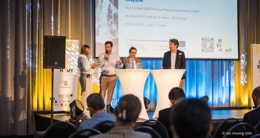 Eine Moderatorin und drei Diskussionsteilnehmer stehen beim Bioeconomy Summit 2026 auf einer B&uuml;hne an zwei wei&szlig;en Stehtischen. Ein Mann spricht in ein Mikrofon. Im Hintergrund zeigt eine Pr&auml;sentationsfolie die Namen der Teilnehmenden und das Logo von Bioeconomy Austria.