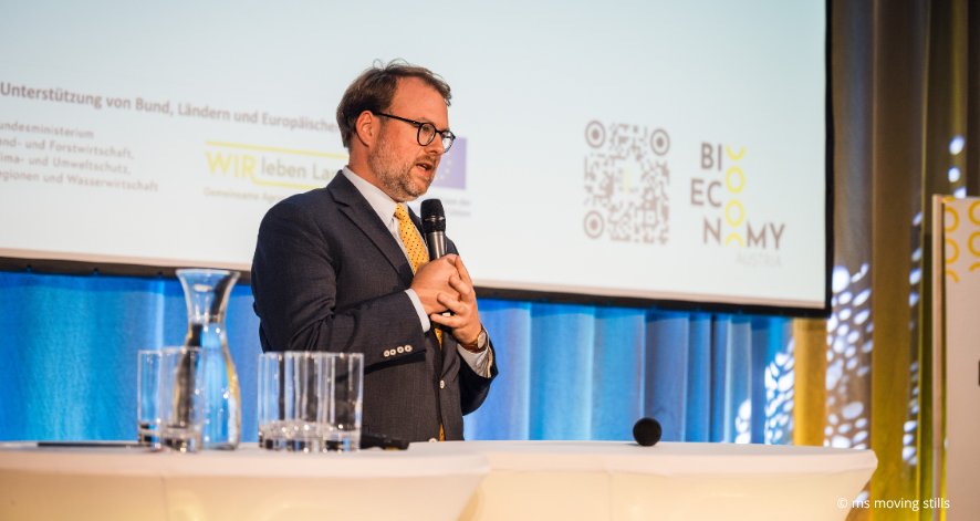 Ein Mann mit Brille, dunklem Anzug und gelber Krawatte spricht beim Bioeconomy Summit 2026 in ein Handmikrofon. Er steht hinter einem wei&szlig;en Stehtisch mit Wassergl&auml;sern und Karaffen. Im Hintergrund sind eine Pr&auml;sentationsfolie mit dem Bioeconomy-Austria-Logo und blaue Vorh&auml;nge zu sehen.