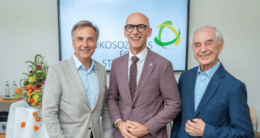 Drei Vertreter des &Ouml;kosozialen Forums Steiermark stehen l&auml;chelnd nebeneinander vor dem Logo der Organisation.