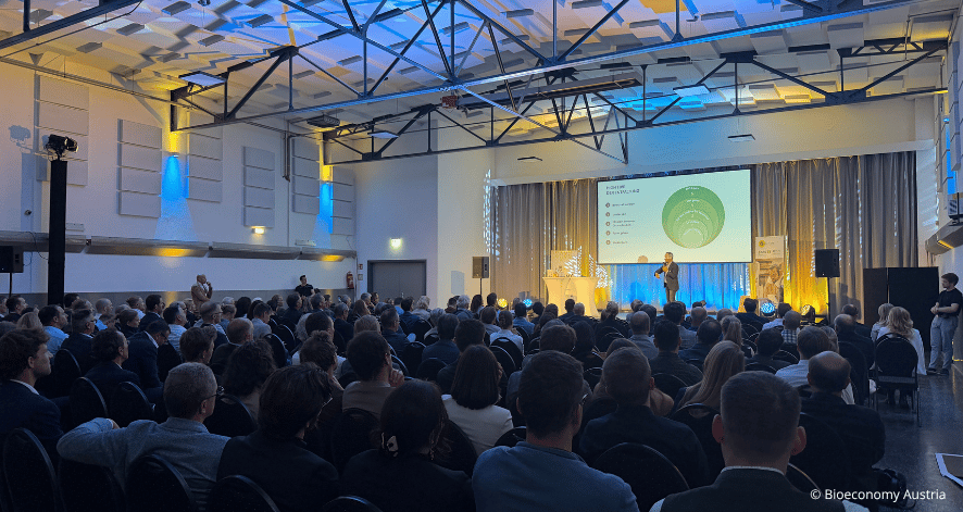 Blick in einen gro&szlig;en, voll besetzten Veranstaltungssaal beim Bioeconomy Summit 2026. Zahlreiche Besucherinnen und Besucher verfolgen einen Vortrag auf der B&uuml;hne. Eine gro&szlig;e Pr&auml;sentationsleinwand zeigt eine Grafik mit gr&uuml;nen Kreisen. Die Halle ist mit blauen und gelben Lichteffekten beleuchtet.
